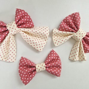 May include: Four fabric bows in different sizes. The bows are made from two fabrics: a dusty rose with white polka dots and a cream fabric with matching polka dots. The bows are tied in the centre with the cream fabric.