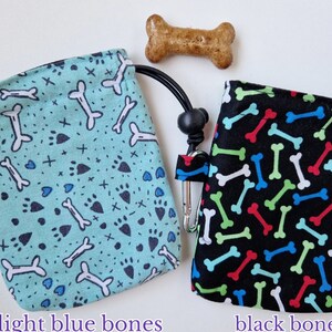 May include: Two fabric treat pouches with drawstring closures and metal clips. One pouch is light blue with a pattern of white bones, black paw prints, and small crosses. The other is black with a pattern of red, green, blue, and white bones.
