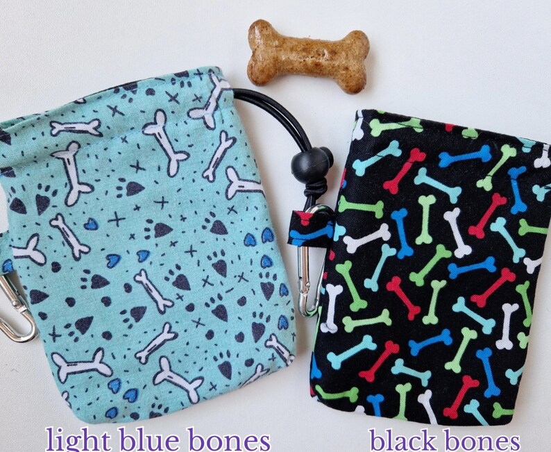 May include: Two fabric treat pouches with drawstring closures and metal clips. One pouch is light blue with a pattern of white bones, black paw prints, and small crosses. The other is black with a pattern of red, green, blue, and white bones.