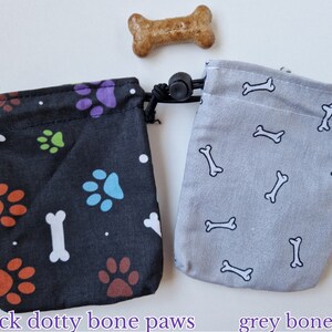 May include: Two small fabric drawstring pouches with a dog treat. The black pouch features colourful paw prints, white bones, and dots. The grey pouch features a repeating pattern of white bones. Text below reads black dotty bone paws and grey bones.