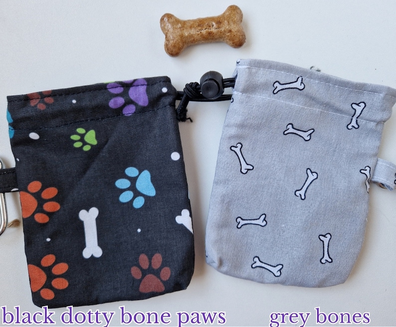 May include: Two small fabric drawstring pouches with a dog treat. The black pouch features colourful paw prints, white bones, and dots. The grey pouch features a repeating pattern of white bones. Text below reads black dotty bone paws and grey bones.