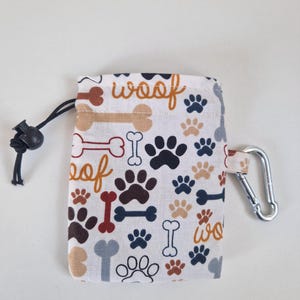 May include: A rectangular fabric pouch featuring a repeating pattern of dog paw prints, bones, and the word woof in shades of brown, red, blue, and grey. The pouch includes a black drawstring closure with a toggle and a silver metal carabiner clip.