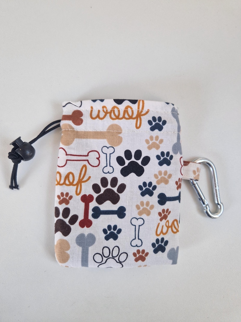 May include: A rectangular fabric pouch featuring a repeating pattern of dog paw prints, bones, and the word woof in shades of brown, red, blue, and grey. The pouch includes a black drawstring closure with a toggle and a silver metal carabiner clip.