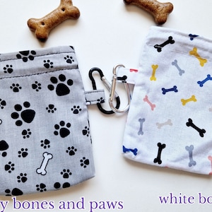 May include: Two rectangular fabric pouches with drawstring closures and metal carabiners. One pouch is grey with a black paw print and white bone pattern, while the other is white with a multi-coloured bone pattern. Text reads grey bones and paws and white bones.
