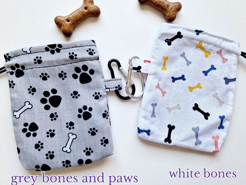 May include: Two rectangular fabric pouches with drawstring closures and metal carabiners. One pouch is grey with a black paw print and white bone pattern, while the other is white with a multi-coloured bone pattern. Text reads grey bones and paws and white bones.