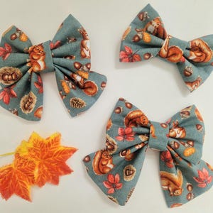 May include: Three fabric hair bows in a muted blue-green colour featuring a repeating pattern of brown squirrels, orange autumn leaves, acorns, and pinecones. The set includes two larger bows and one smaller bow, accompanied by two orange maple leaves.