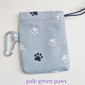 May include: A light green rectangular fabric pouch featuring a repeating pattern of dark navy blue and white paw prints. The pouch includes a silver metal carabiner attached to the side and a black drawstring cord with a plastic toggle at the top.