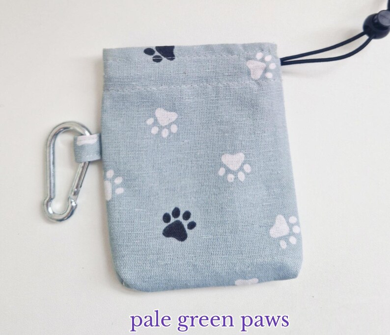 May include: A light green rectangular fabric pouch featuring a repeating pattern of dark navy blue and white paw prints. The pouch includes a silver metal carabiner attached to the side and a black drawstring cord with a plastic toggle at the top.