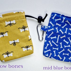 May include: Two rectangular fabric treat pouches with drawstring closures and metal carabiners. The left pouch is mustard yellow with a white bone and black bow pattern. The right pouch is blue with a white bone pattern. Text reads yellow bones and mid blue bones.