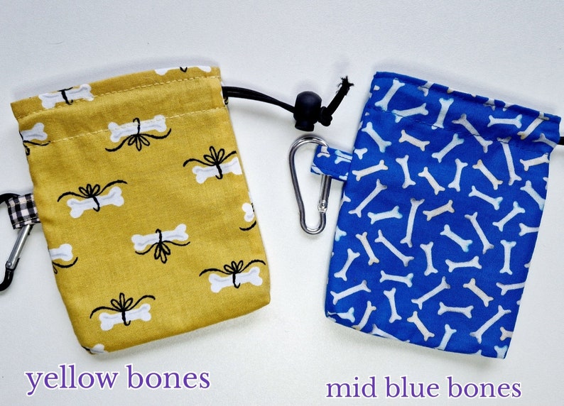 May include: Two rectangular fabric treat pouches with drawstring closures and metal carabiners. The left pouch is mustard yellow with a white bone and black bow pattern. The right pouch is blue with a white bone pattern. Text reads yellow bones and mid blue bones.