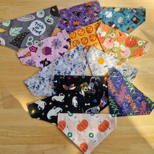 May include: A collection of triangular fabric pet bandanas featuring various Halloween-themed patterns including pumpkins, ghosts, skeletons, spider webs, witch hats, and the text trick or treat and spooky, arranged in a scattered overlapping display.