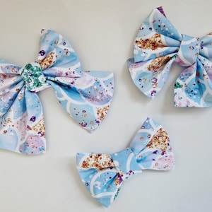 May include: Three blue and white fabric hair bows with a mermaid scale pattern and glitter accents. The bows are different sizes.