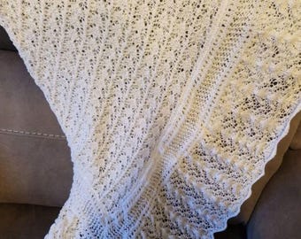 May include: A white, open-weave knitted shawl with a delicate, lacy pattern. The shawl is draped over a surface, showcasing its intricate design and scalloped edges. The soft, off-white color suggests a cozy, handmade item.