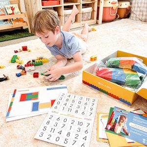 May include: A child plays with colorful plastic building blocks on a white rug. The blocks are in a yellow box with the text "@yay_a_play" printed on the side. There are also several worksheets with numbers and patterns on them.