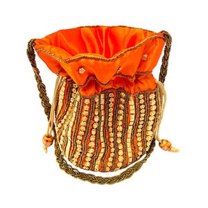 May include: An orange drawstring bag with a beaded design. The bag features a velvet-like orange top and a body adorned with vertical rows of gold and white beads. The handles are made of twisted brown beads, and the bag is set against a white background.