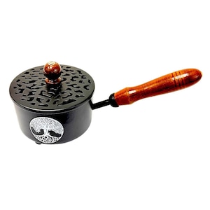 May include: A black metal incense burner with a long wooden handle and a round lid featuring a cutout pattern. The side of the burner displays a white circular emblem of a tree with visible roots. A small wooden knob sits atop the lid.