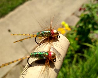 May include: Two green and gold fly fishing lures with red accents are attached to a cork. The lures have brown and yellow feathers.