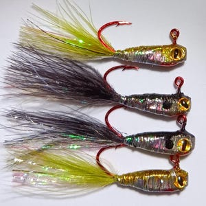 May include: Four fishing lures with black, green, and gold colored feathers and a silver body. The lures have a red hook and a gold eye.
