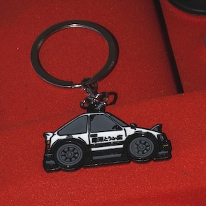 May include: A black and white car-shaped keychain with a silver key ring. The keychain has a detailed design of a car with black wheels and text on the side. The background is a textured red surface.
