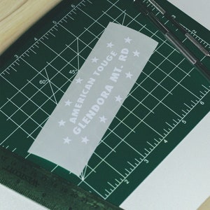 May include: A white vinyl sticker with the text "AMERICAN TOUGE GLENDORA MT. RD" and stars around the text. The sticker is on a green cutting mat.