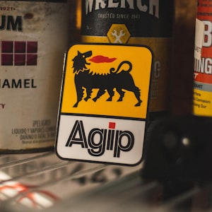 Könnte beinhalten: Ein quadratischer Aufkleber mit dem Agip-Logo, das ein schwarzes, hundeähnliches Wesen mit roter, flammenartiger Zunge auf gelbem Hintergrund zeigt. Das Wort "Agip" ist darunter in Schwarz gedruckt.