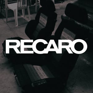 May include: Two black automotive bucket seats featuring the RECARO brand name embroidered in white on the headrests and backrests. The seats have adjustable side bolsters and integrated headrests, with the RECARO logo prominently displayed.