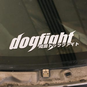 May include: A white vinyl decal applied to a dark glass surface. The decal features the word dogfight in a stylised, slanted font, with the Japanese text 成田ドックファイト positioned directly underneath the right side of the main lettering.