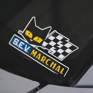 May include: A sticker featuring a black cat with yellow eyes and a checkered flag design. The sticker has the text "S.E.V. MARCHAL" in blue and yellow. The sticker is on a black surface.