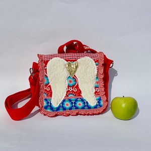 Könnte beinhalten: Rote und weiße karierte Stofftasche mit einem weißen und goldenen Engelsflügel-Design. Die Tasche hat einen roten Trageriemen und einen Rüschenrand.