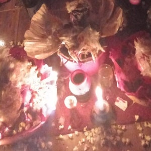 Puede incluir: Un altar de tela roja con una variedad de artículos, incluyendo velas, plumas y un tazón de líquido rojo. El altar está decorado con plumas blancas y palomitas de maíz.