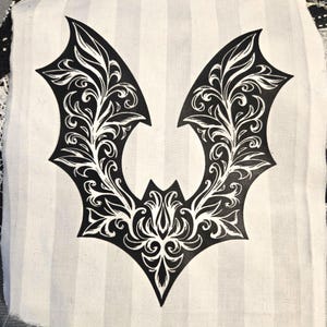 Könnte beinhalten: Ein schwarz-weißes fledermausförmiges Design mit aufwendigen floralen Details. Das Design ist auf einem weißen Stoff mit vertikalen grauen Streifen gedruckt. Die Flügel der Fledermaus sind ausgebreitet und das Design ist symmetrisch.