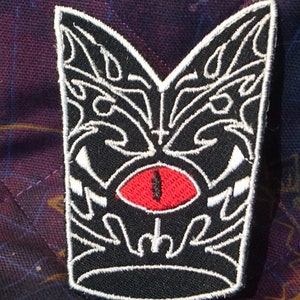 May include: Black and white embroidered patch with a red eye in the center. The patch features a stylized design of a creature's face with sharp teeth.