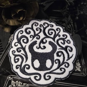 May include: White embroidered patch with a black silhouette of a creature with swirling vines around it.