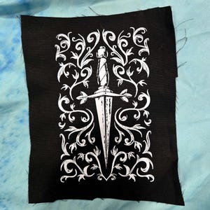 Op de afbeelding: Zwarte patch met een witte illustratie van een dolk, met een decoratieve handgreep en pareerstang, omringd door wervelende wijnranken en bladeren. Het ontwerp is gecentreerd.