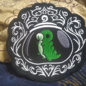 May include: Embroidered patch with a green creature in a glass orb on a black background. The patch has a decorative white border with swirling designs. The creature is holding a smaller, light green figure.