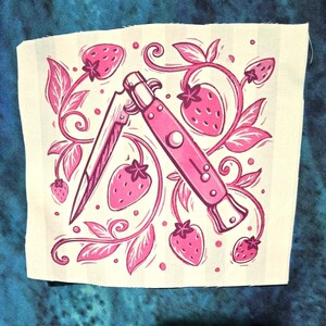 Könnte beinhalten: Ein quadratischer Stoffaufnäher mit einer rosa-weißen grafischen Illustration eines Klappmessers, umgeben von Erdbeerranken, Blättern und Beeren. Das Motiv ist auf einem hellcremefarbenen Hintergrund mit dezenten vertikalen Streifen gedruckt.