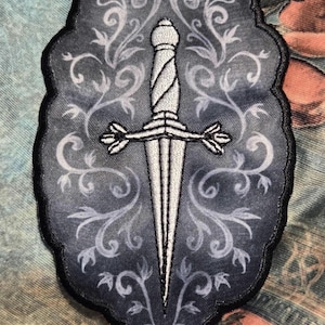 Pode incluir: Patch bordado com uma adaga prateada com guarda e punho detalhados, sobre um fundo cinza com um desenho floral rodopiante. O patch tem uma borda preta. Ideal para jaquetas, bolsas ou outros artigos de tecido.