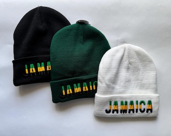 May include: Three knit beanies in black, green, and white. Each beanie features the word "Jamaica" embroidered in yellow, green, and black on the folded brim. The beanies are displayed on a white surface.