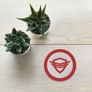 May include: A round, embroidered patch with a red border and a white center. The center features a red shield design with wings. Two small potted succulents sit on a light wood surface.