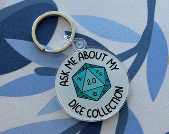 May include: A circular acrylic keychain featuring a light blue twenty-sided die illustration in the center. The text "ASK ME ABOUT MY DICE COLLECTION" is printed in black capital letters around the die. A silver metal split ring is attached.