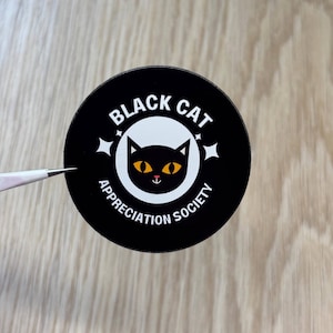 Pode incluir: Um autocolante redondo preto com o texto "BLACK CAT APPRECIATION SOCIETY" em branco. O autocolante apresenta um gato preto com olhos amarelos e nariz rosa. Duas estrelas brancas estão em ambos os lados do gato.