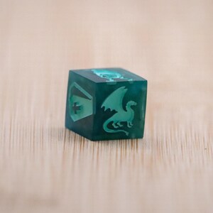 Puede incluir: Un dado de seis caras de color verde azulado con diseños grabados. Un lado presenta un dragón, otro un escudo con una cruz. El dado está hecho de un material translúcido, y los diseños son de un tono verde azulado más claro. El dado está sobre una superficie de madera marrón claro.
