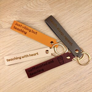 May include: Four rectangular faux leather keychains in orange, cream, dark grey, and maroon. Each features a gold-tone metal rivet and keyring. The orange tag reads "just vibing but teaching," cream reads "teaching with heart," grey reads "teach, sleep, repeat," and maroon reads "caffeinated and teaching" with a coffee cup icon.