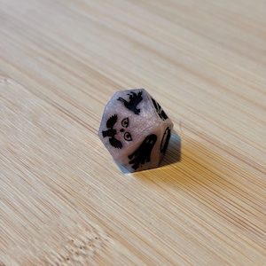 Puede incluir: Un dado de doce caras con un acabado nacarado e ilustraciones negras de un fantasma, un búho y murciélagos. El dado está sobre una superficie de madera.