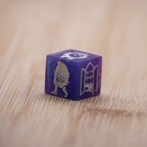 Puede incluir: Un dado morado de seis caras con diseños grabados en color plata. El dado presenta una figura caminando, un monstruo en una puerta y otros símbolos. El dado está sobre una superficie de madera marrón claro.