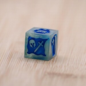 Puede incluir: Un dado azul translúcido en forma de cubo con símbolos azul oscuro. Los símbolos incluyen una calavera, una espada y un escudo, y un ataúd. Este dado se utiliza probablemente para juegos de mesa o juegos de rol.
