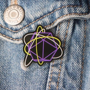 May include: A black enamel pin with a geometric design in purple, yellow, and white, attached to a denim jacket. The pin features a complex shape with intersecting lines and a white outline, set against the blue denim.