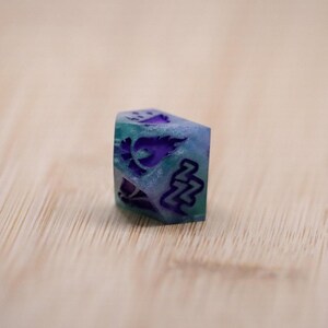 Puede incluir: Un dado de seis caras hecho a mano con una base azul-verdosa translúcida y diseños morados grabados. El dado presenta símbolos y patrones, incluido un diseño floral y un símbolo estilizado 'NN'. Se encuentra sobre una superficie de madera marrón claro.