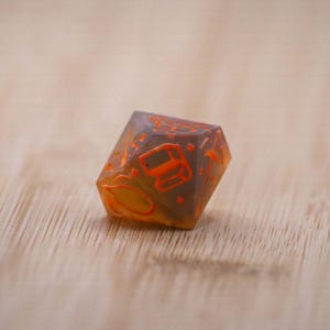 May include: A multi-sided, translucent die with orange internal designs. The die has a gray and orange color scheme, with orange symbols and shapes visible inside. The die is resting on a light brown wooden surface.