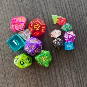 May include: A collection of colorful polyhedral dice in various shapes and sizes. The dice are in shades of pink, red, green, purple, blue, and black, with gold or white numbering. The dice are for tabletop games.